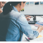 Two Asian women sit at a white desk, reviewing print marketing materials. One holds a photo while the other looks thoughtful. A computer monitor, camera, pens, and scattered prints are visible on the desk.