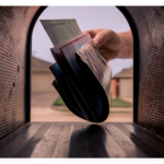 View from inside a mailbox as a hand inserts a stack of print marketing mailpieces. Blurred houses are visible in the background through the open mailbox door.
